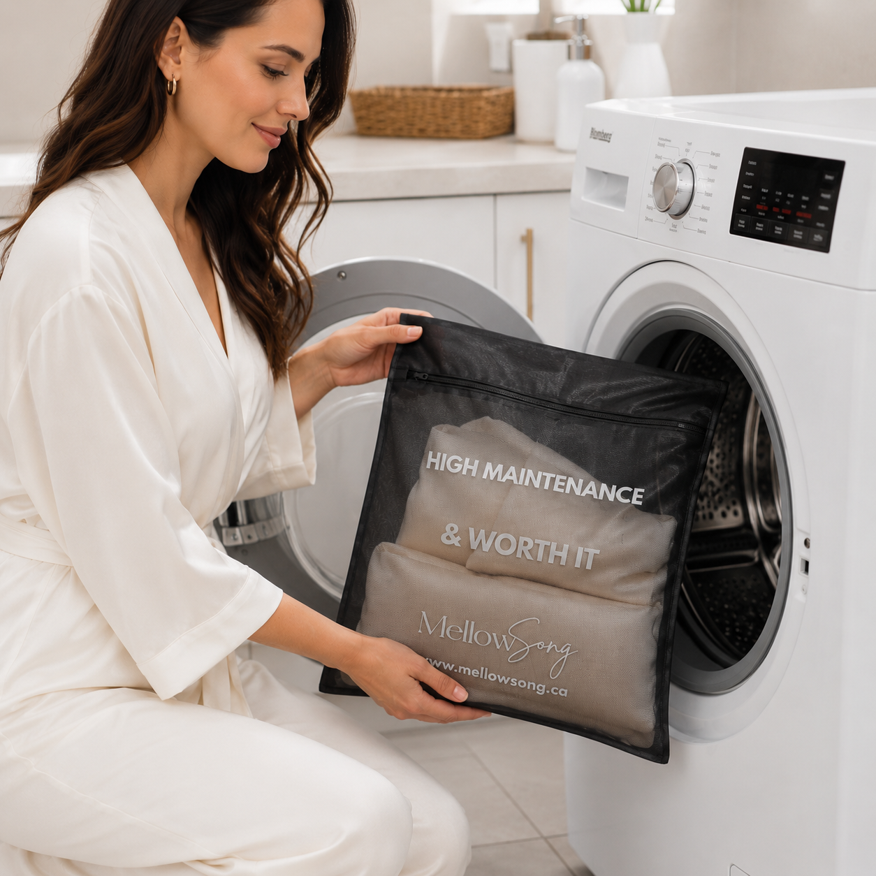 Lifestyle image of a woman loading a Mellow Song black mesh delicates laundry bag into a front-load washing machine. The bag reads "HIGH MAINTENANCE & WORTH IT" and holds silk sleep accessories.