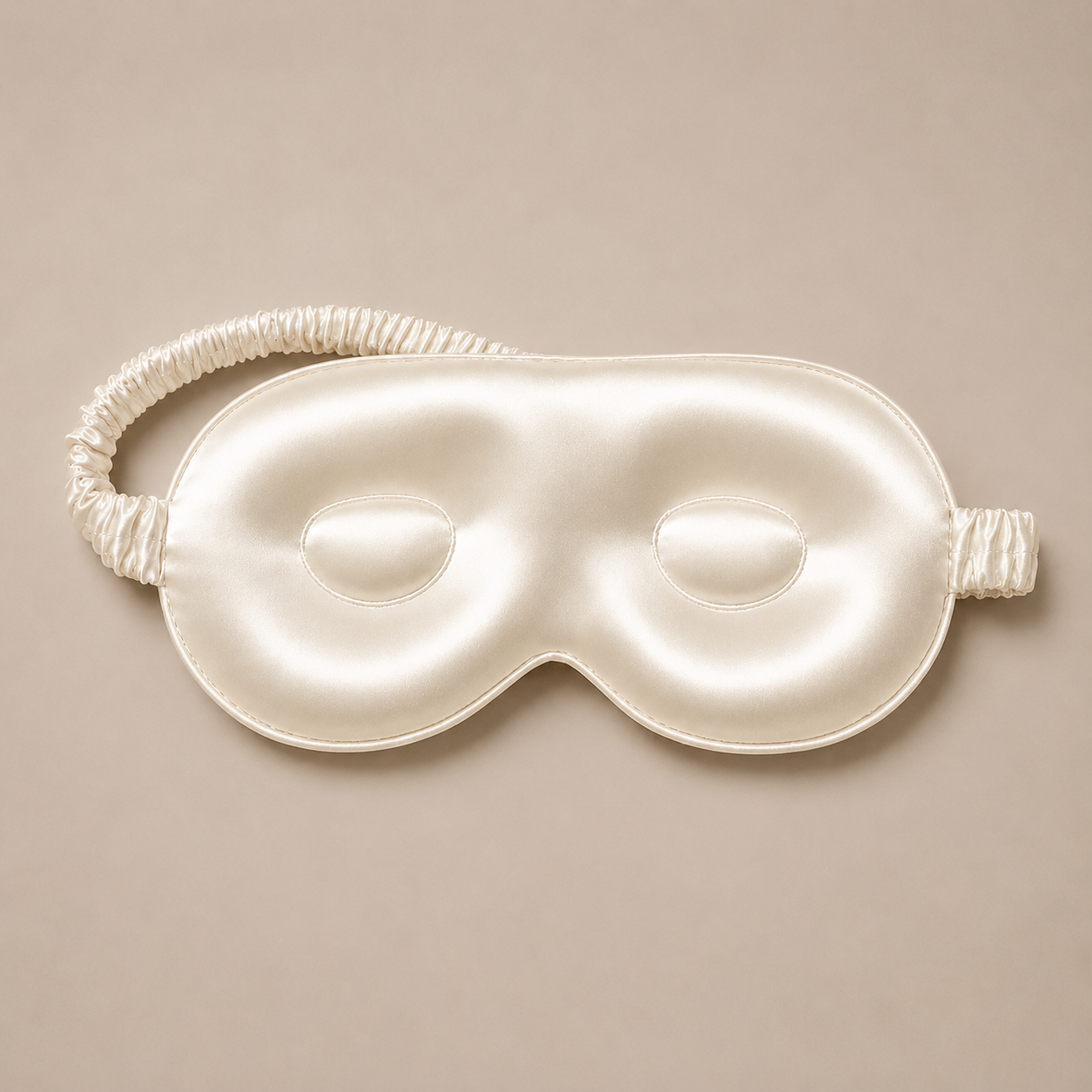 Cream-colored silk sleep mask with a matching ruched elastic headband, centered against a neutral beige background.