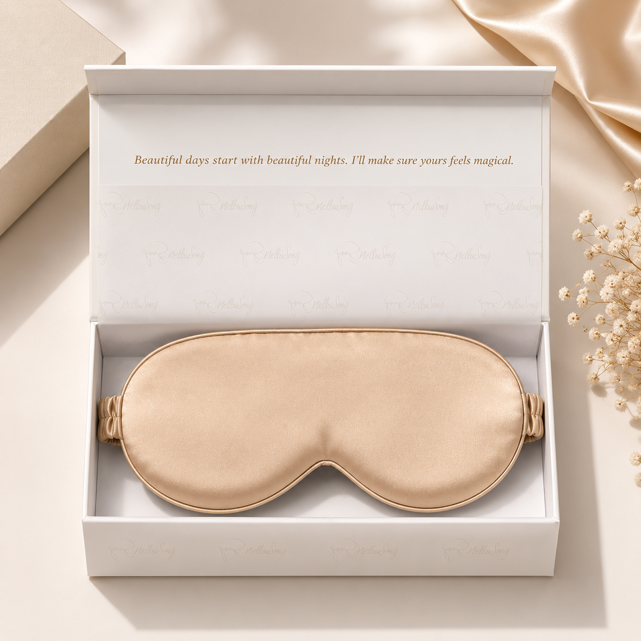 Champagne gold silk eye mask presented in an elegant white branded gift box, styled with silk fabric and baby's breath flowers on a neutral surface.