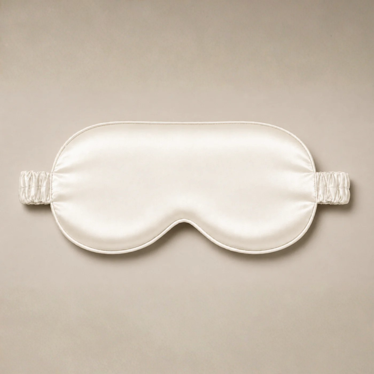 Cream-colored silk sleep mask with a matching ruched elastic headband, centered against a neutral beige background.