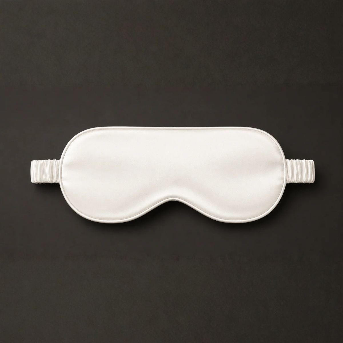 Off-white silk sleep mask with a matching ruched elastic strap, isolated against a dark gray background.